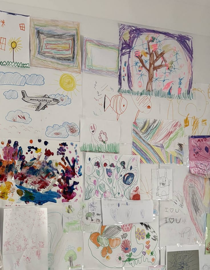 A wall in a family home covered with a child’s drawings — rainbows, a plane, a sun, a tree, painted handprints.