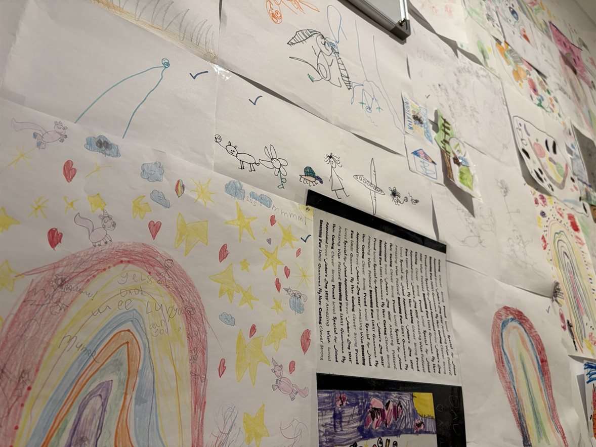 An angled view along a lived-in office wall covered with a child’s colourful drawings, including a large rainbow in the foreground.